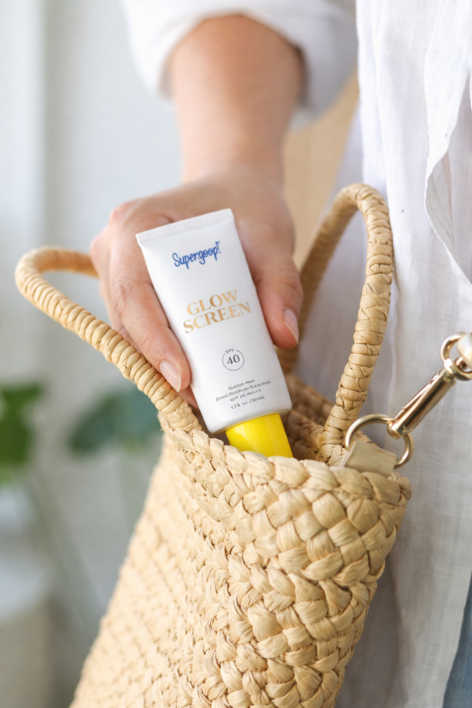 Woman placing Supergoop Glow Screen SPF 40 sunscreen into a woven straw bag in soft natural lighting